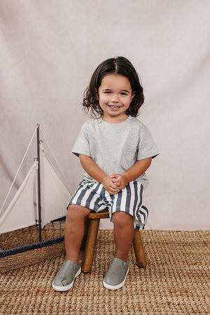 A smiling child sits on a wooden stool, wearing a grey pocket t-shirt and striped shorts, with a sailboat decoration.