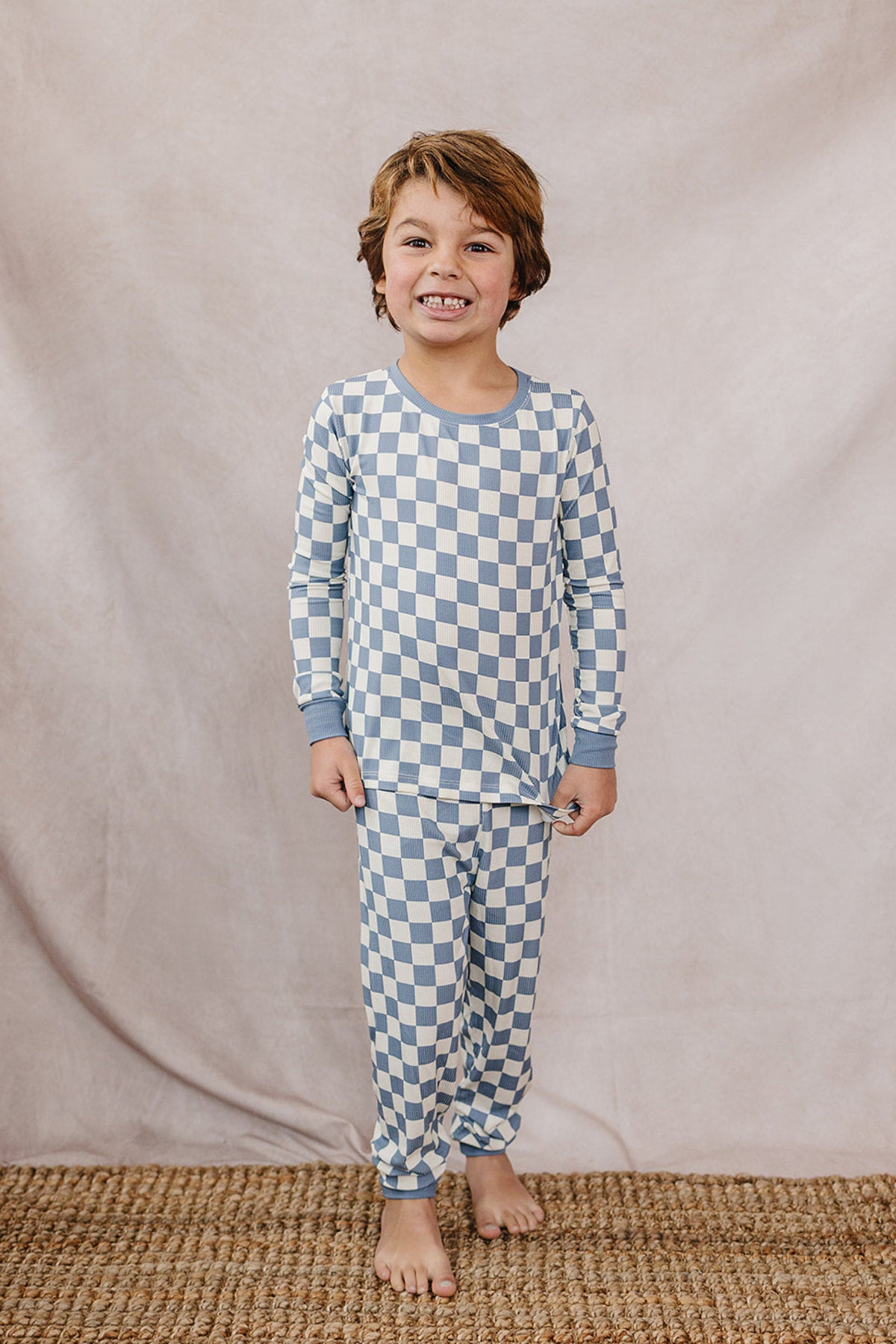 A child smiles while wearing a blue checkered ribbed bamboo cozy set, standing on a woven rug.