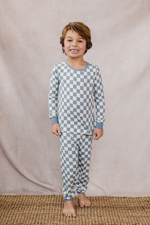 A child smiles while wearing a blue checkered ribbed bamboo cozy set, standing on a woven rug.
