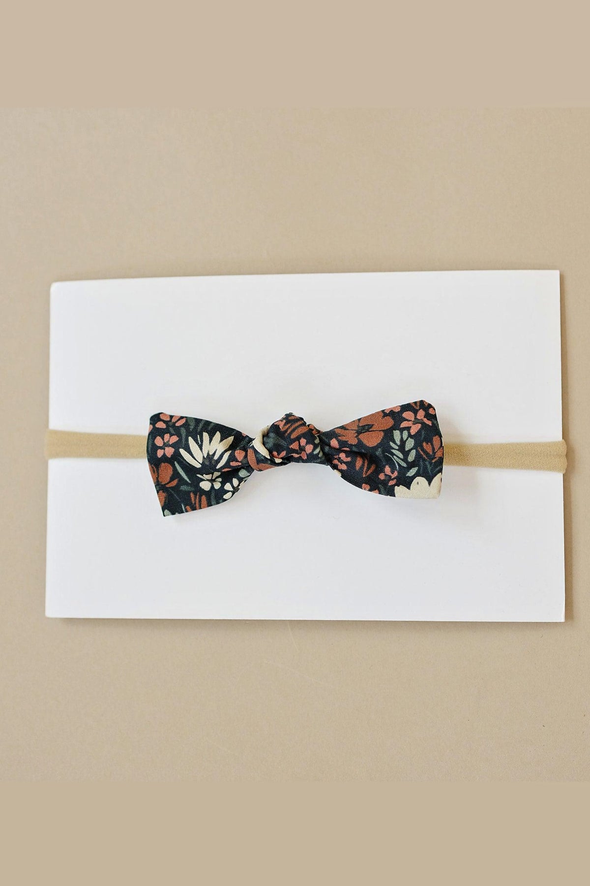 A floral-patterned fabric bow on a beige elastic headband, displayed on a white card against a tan background.