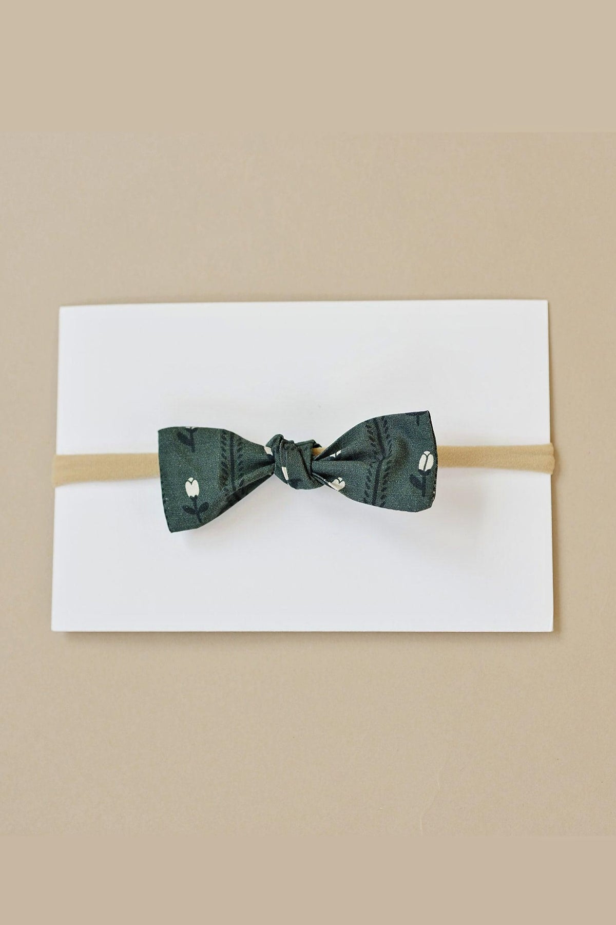A dark green fabric hair bow with a floral pattern on a beige elastic band, displayed on white card.