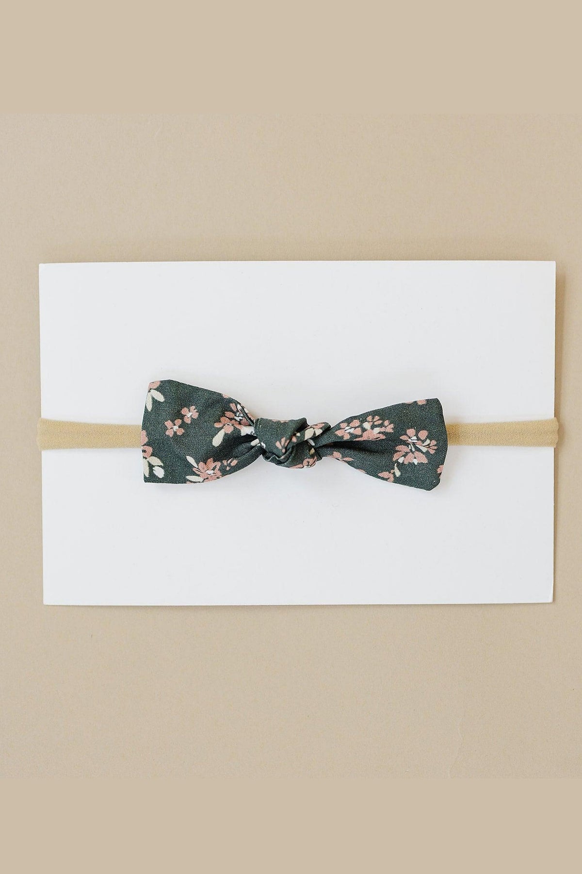 A floral-patterned fabric bow on a beige elastic headband, displayed on a white card background.