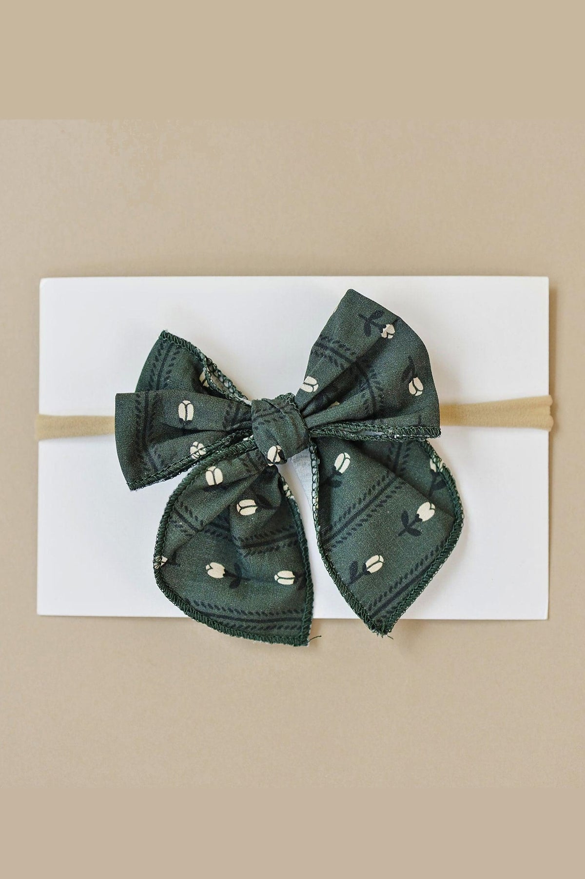 Green fabric hair bow with white floral pattern on a beige elastic band, displayed on a white card.