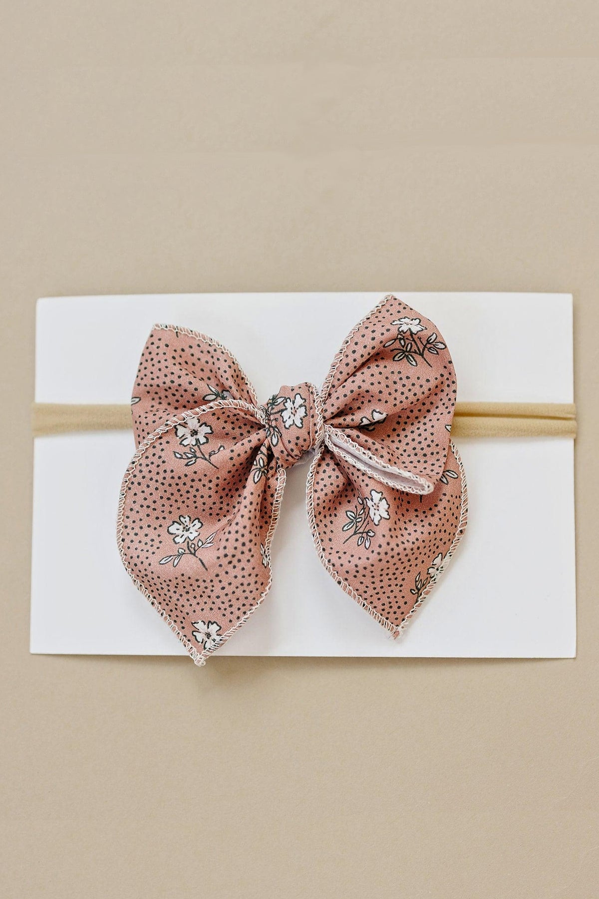 A pink floral-patterned cotton bow with white dots, displayed on a white card with a beige background.