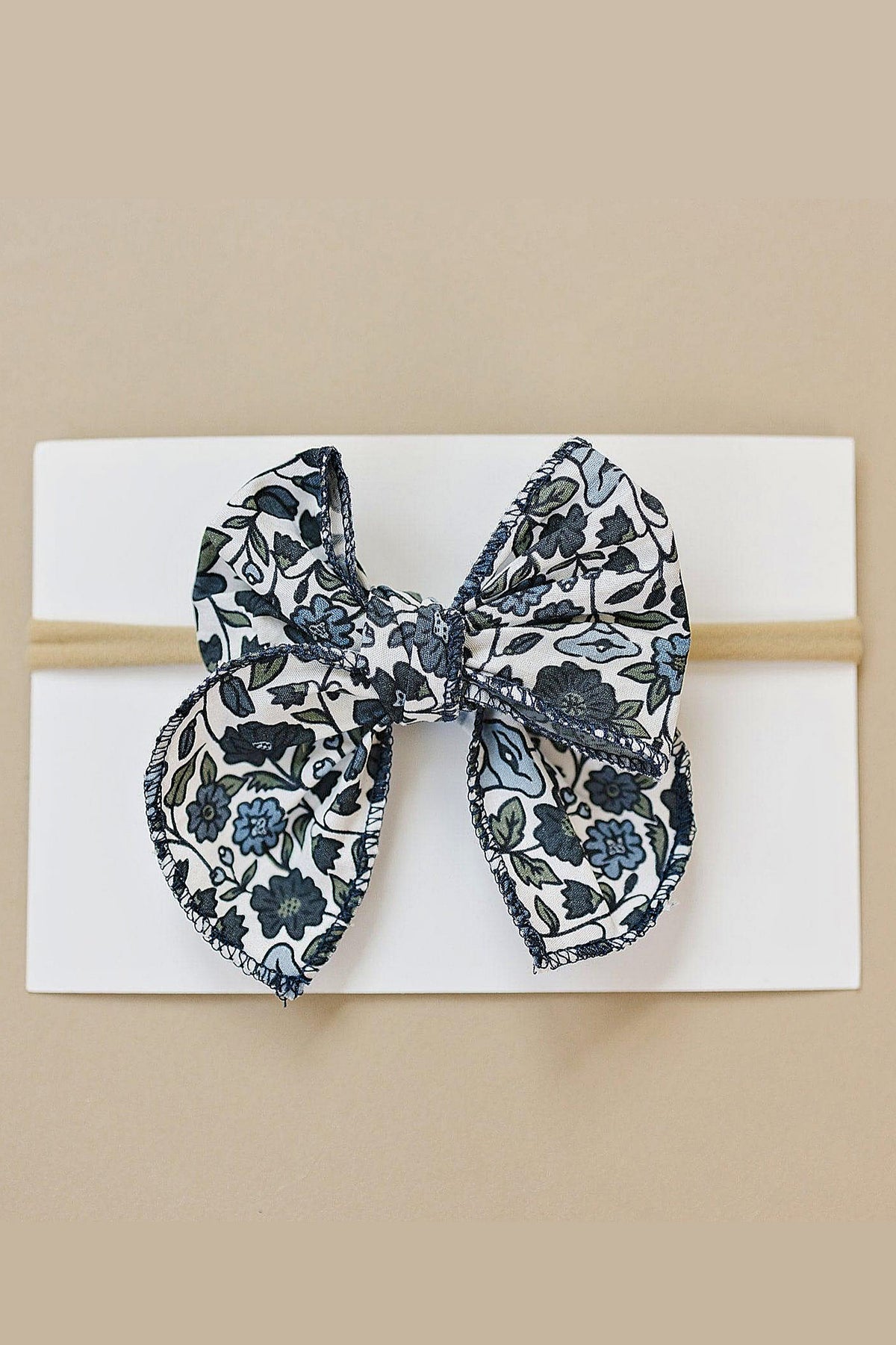 A floral-patterned fabric bow with blue and green hues displayed on a white card against a beige background.