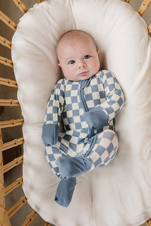 A baby in blue checkered bamboo pajamas rests in a white cushioned wicker basket.