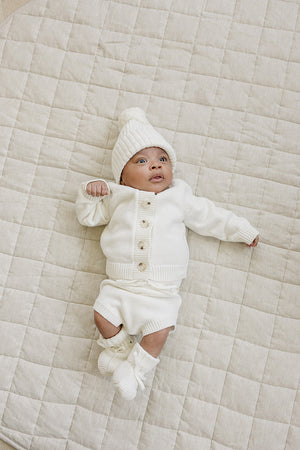 A baby wearing a cozy white knit outfit, including a button-up cardigan, shorts, and matching booties, lies on a textured cream-colored blanket, showcasing a charming and comfortable look.