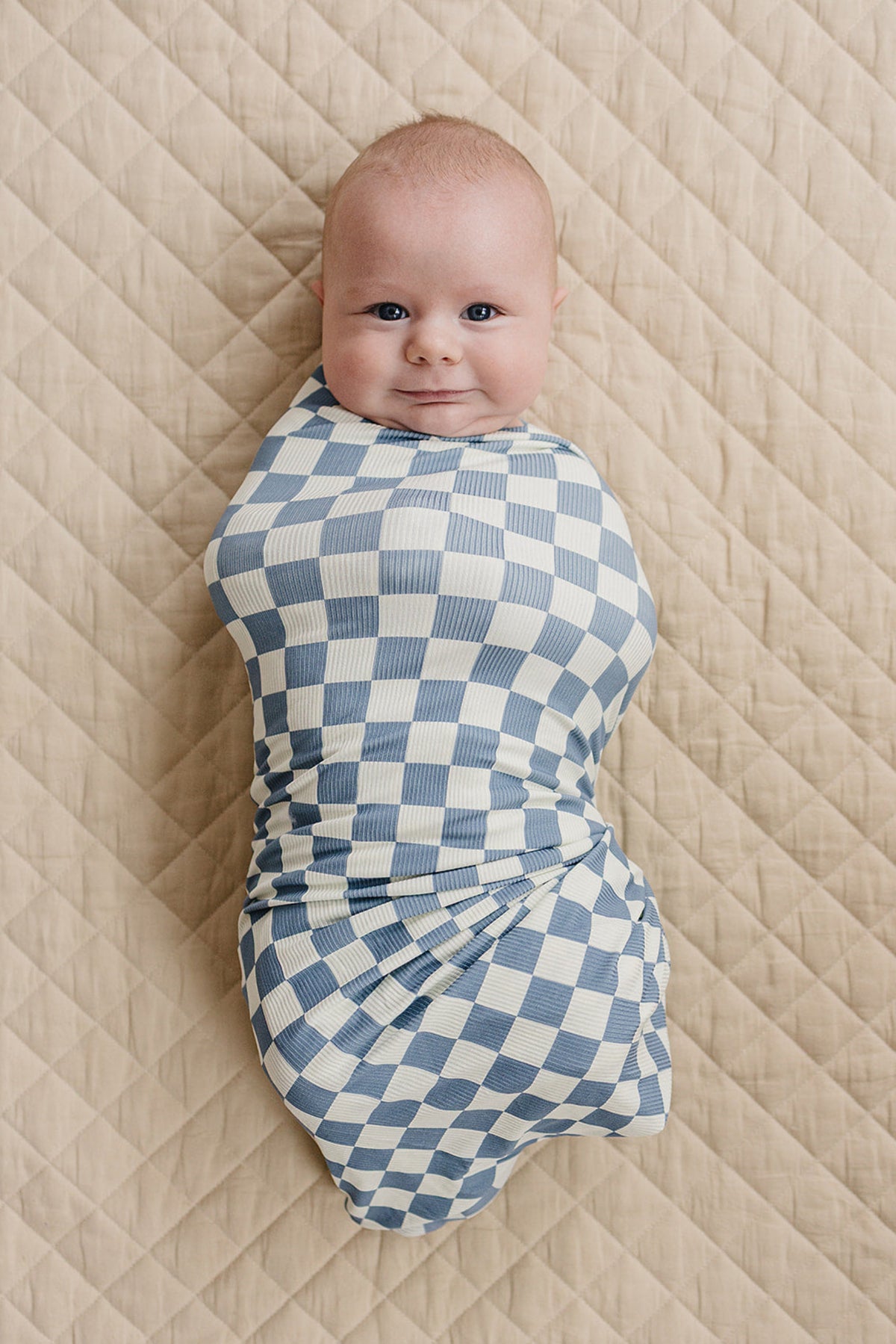 A baby wrapped in a blue checkered swaddle lies on a quilted beige blanket, smiling contentedly.