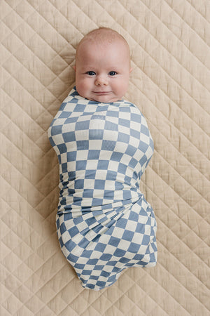 A baby wrapped in a blue checkered swaddle lies on a quilted beige blanket, smiling contentedly.