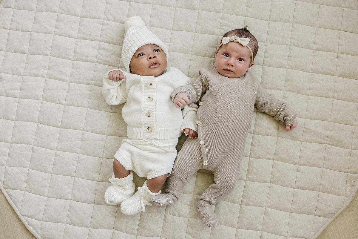 Two infants lie on a beige quilted blanket, one wearing a white knit outfit with a matching hat and booties, while the other is dressed in a soft beige onesie.