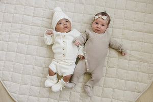 Two infants lie on a quilted beige blanket, one wearing a cream knit sweater and bloomers with a matching hat, and the other in a light taupe onesie with buttons.