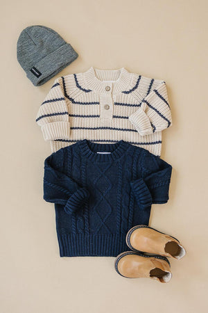 Navy cable knit sweater, cream striped sweater, gray beanie, and tan boots arranged on a beige background.