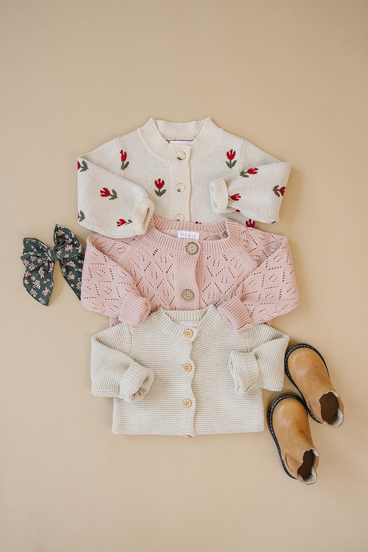 Three knit cardigans in cream, pink, and floral patterns are displayed with a floral bow and tan boots.