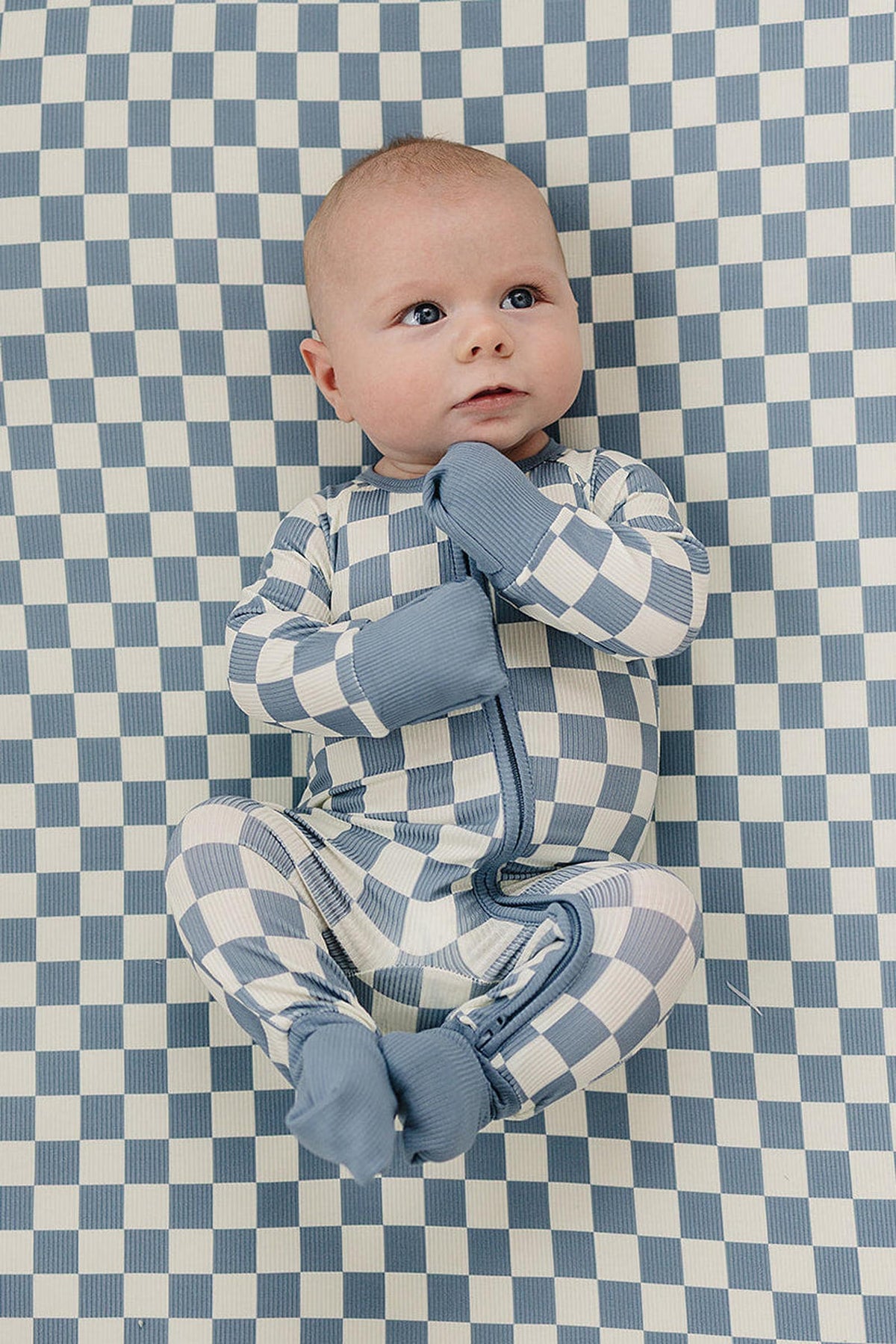 A baby in blue checkered pajamas lies on a matching checkered crib sheet, creating a cohesive, stylish look.