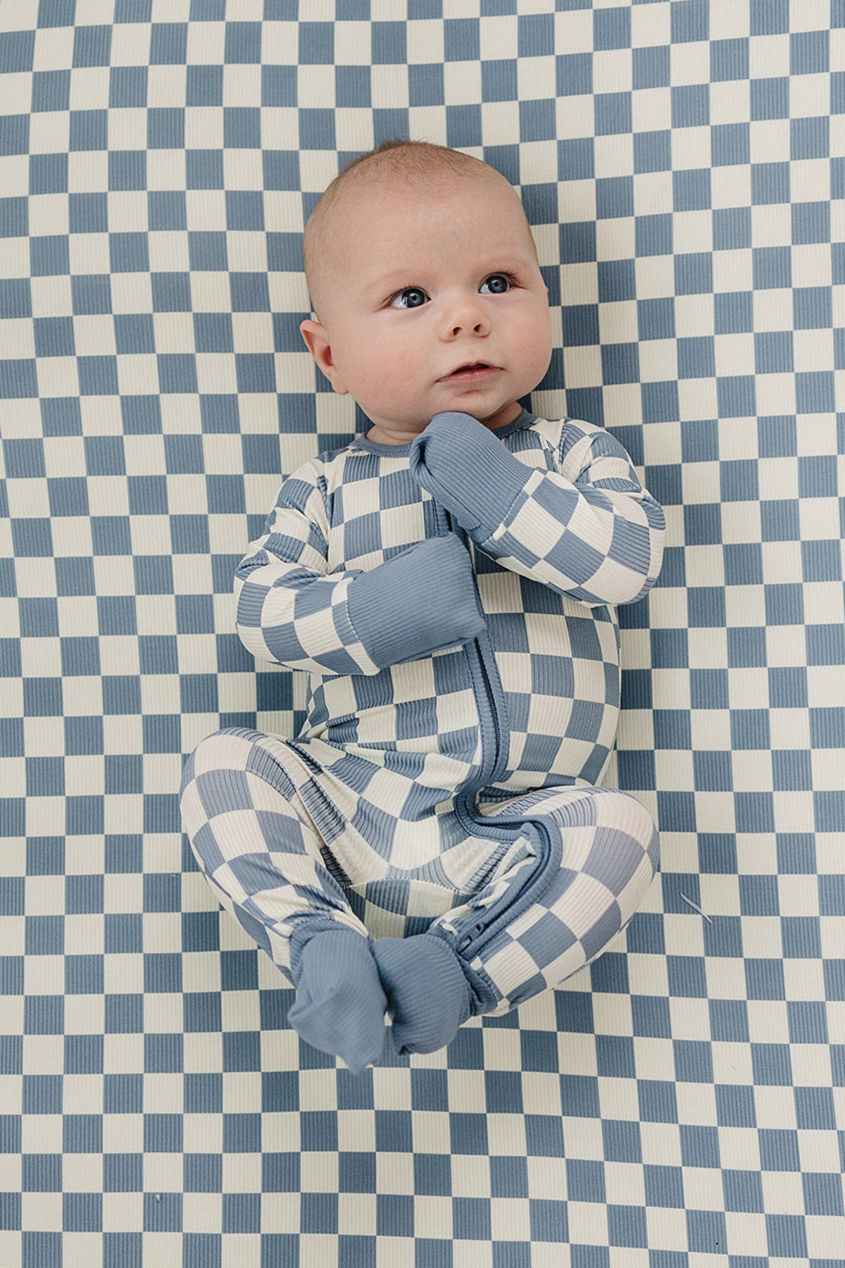 A baby in blue checkered bamboo pajamas lies on a matching checkered crib sheet, looking up.