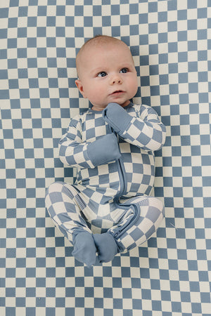 A baby in blue checkered bamboo pajamas lies on a matching checkered crib sheet, looking up.