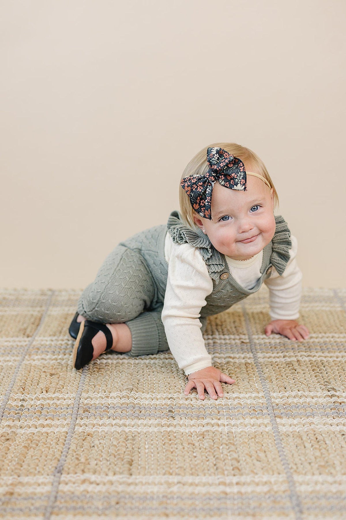 A baby wearing a floral bow headband and green knit overalls crawls on a woven beige rug.