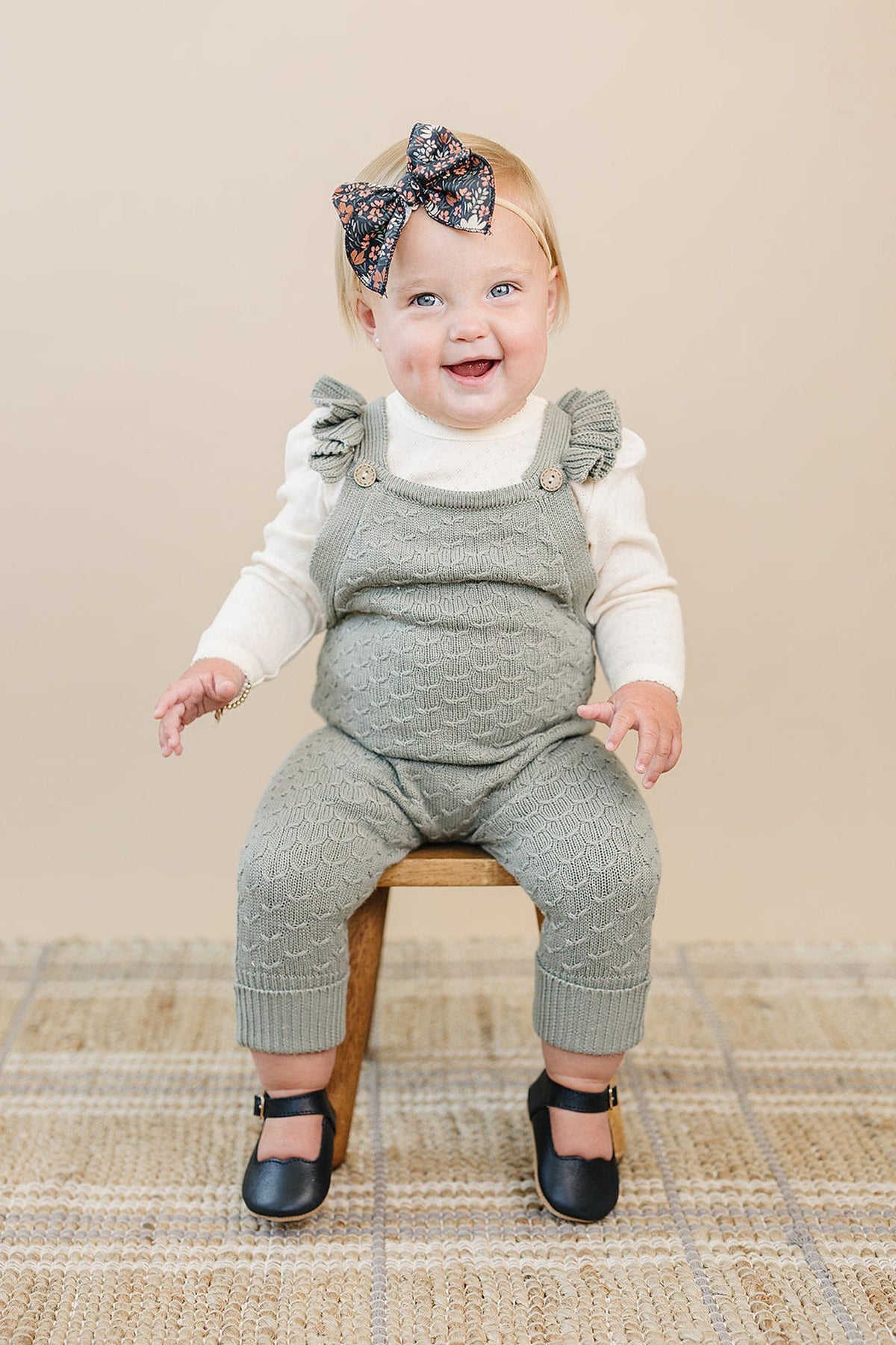 A smiling baby in green ruffle knit overalls and a floral headband sits on a wooden stool.