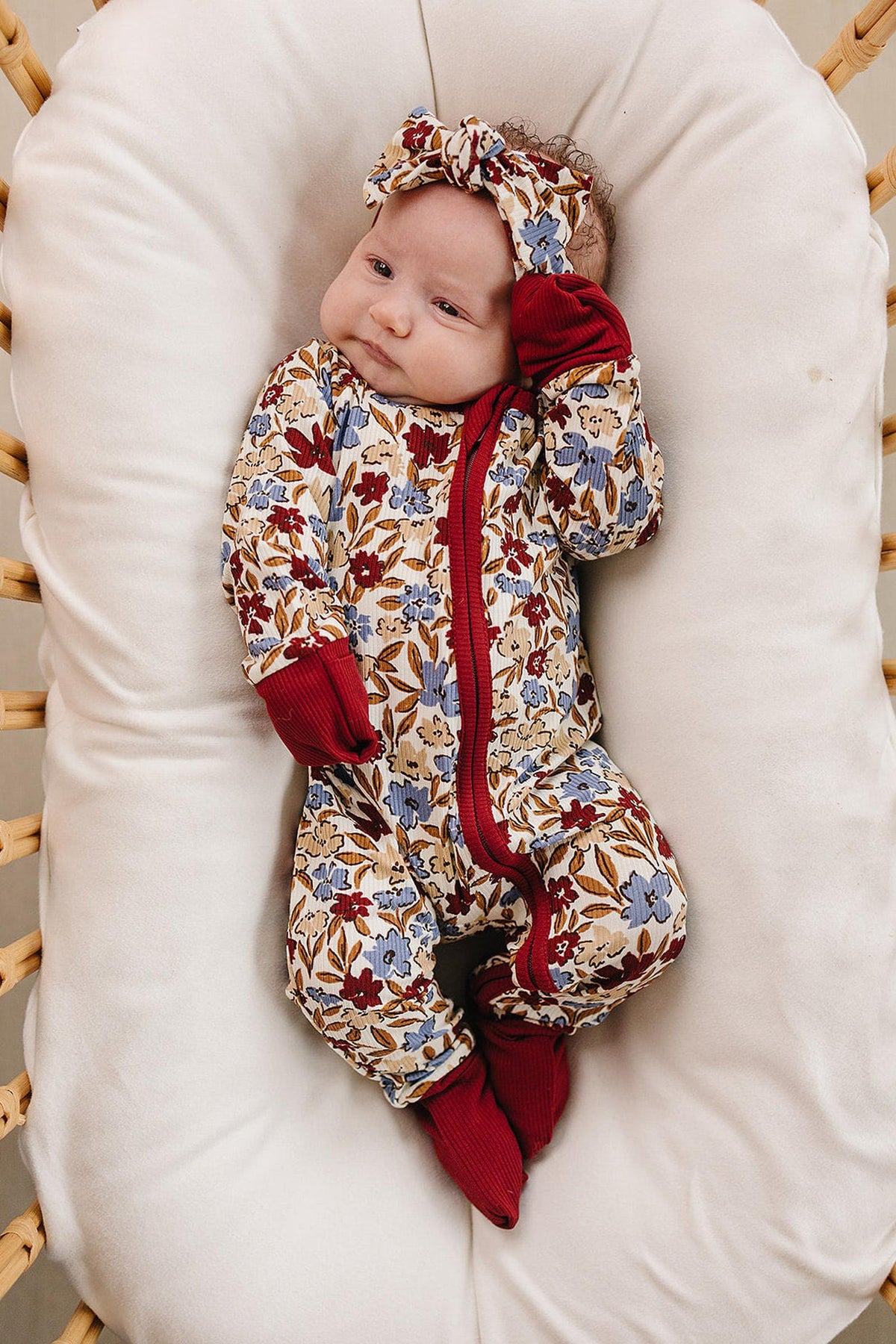 A baby wearing floral bamboo pajamas with a matching headband, lying on a white cushion in a wicker bassinet.