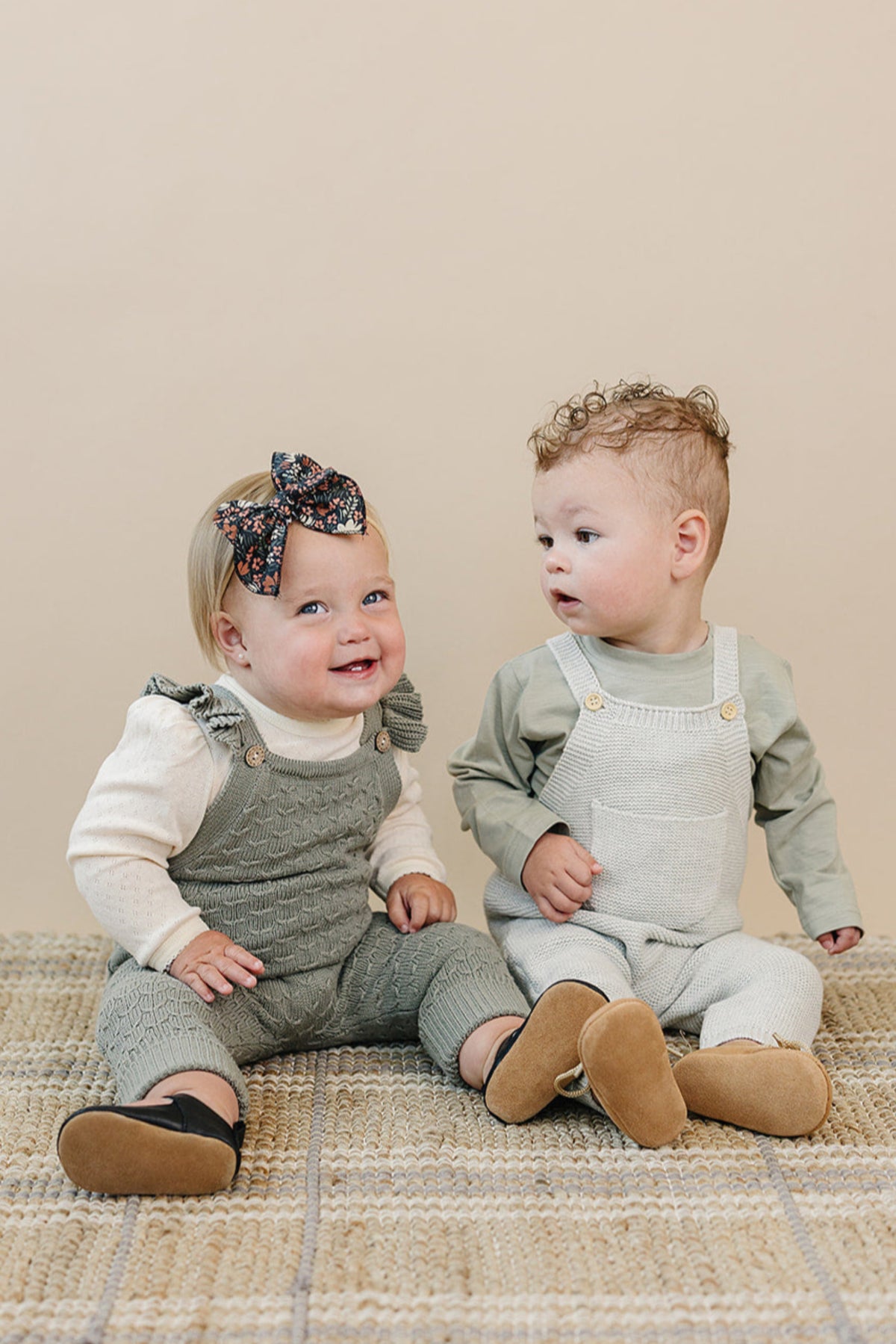 Two toddlers sit on a woven rug wearing knit overalls; one in green with ruffles, the other in light gray.
