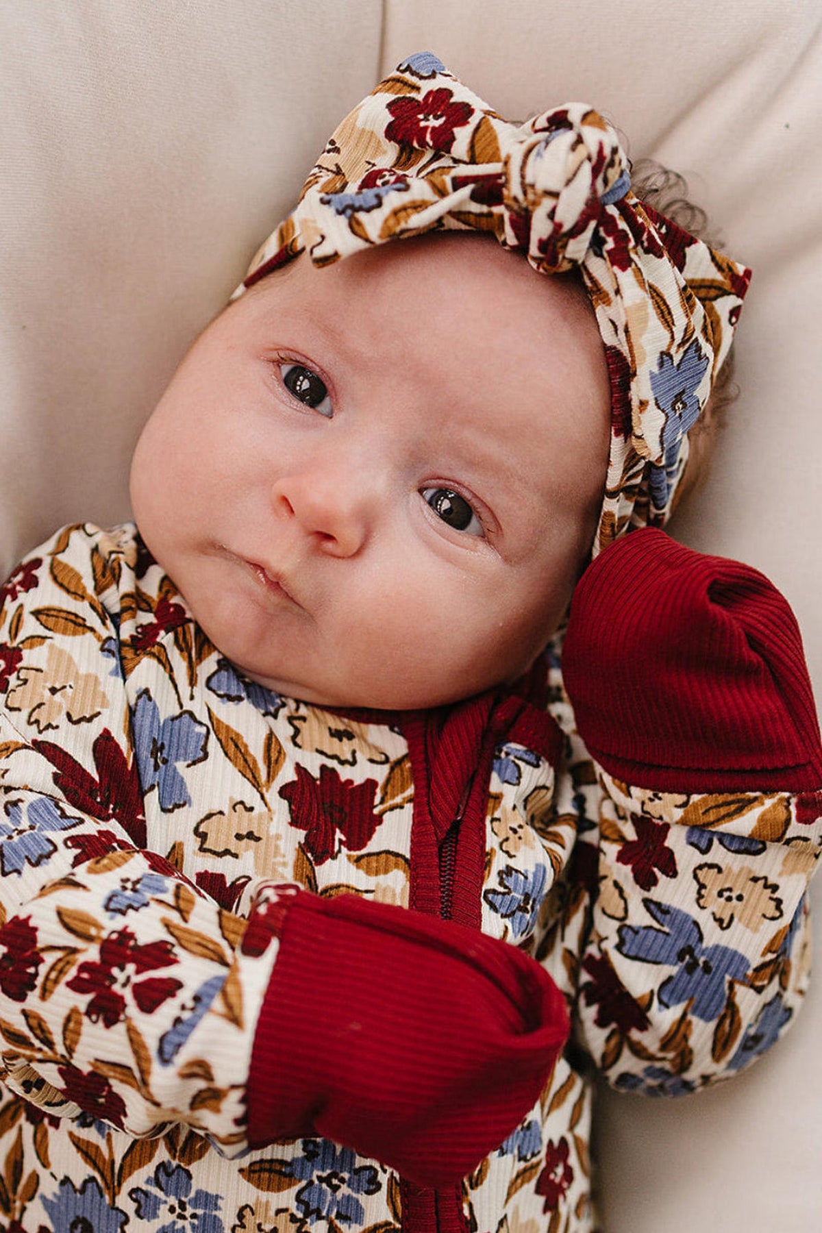 A baby wearing a floral-patterned ribbed bamboo head wrap and matching outfit with red cuffs lies on a cushion.