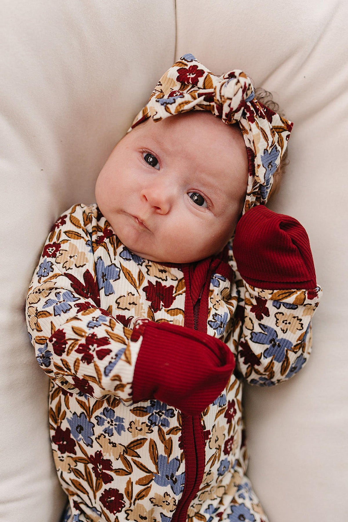 A baby wearing floral-patterned bamboo pajamas with a matching headband and red cuffs, lying on a soft cushion.