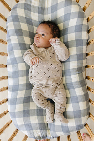 A cozy baby lies on a checkered blanket, wearing a taupe pointelle knit outfit with wooden buttons. The setting features a gentle, soft aesthetic suitable for infants.