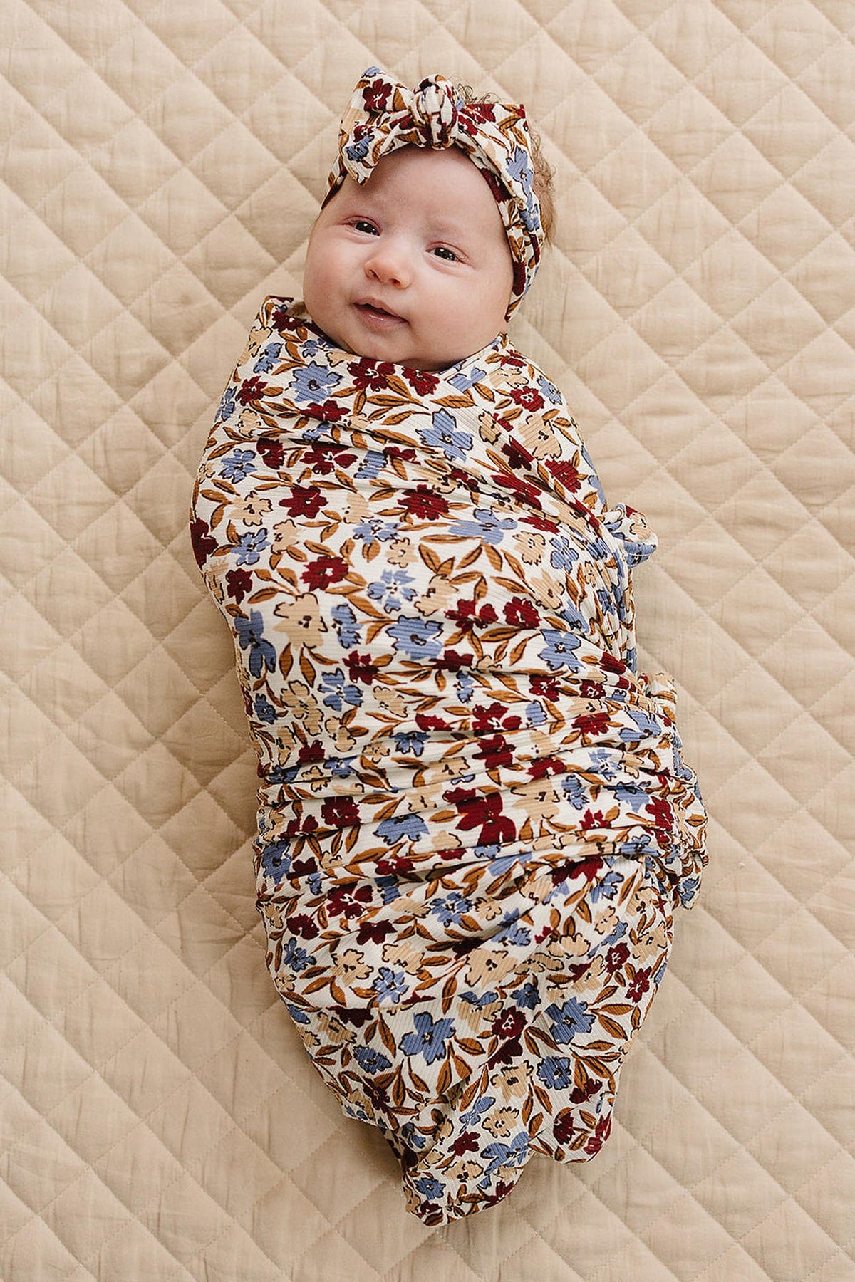 A baby is swaddled in a floral-patterned fabric with matching headband, lying on a quilted beige surface.