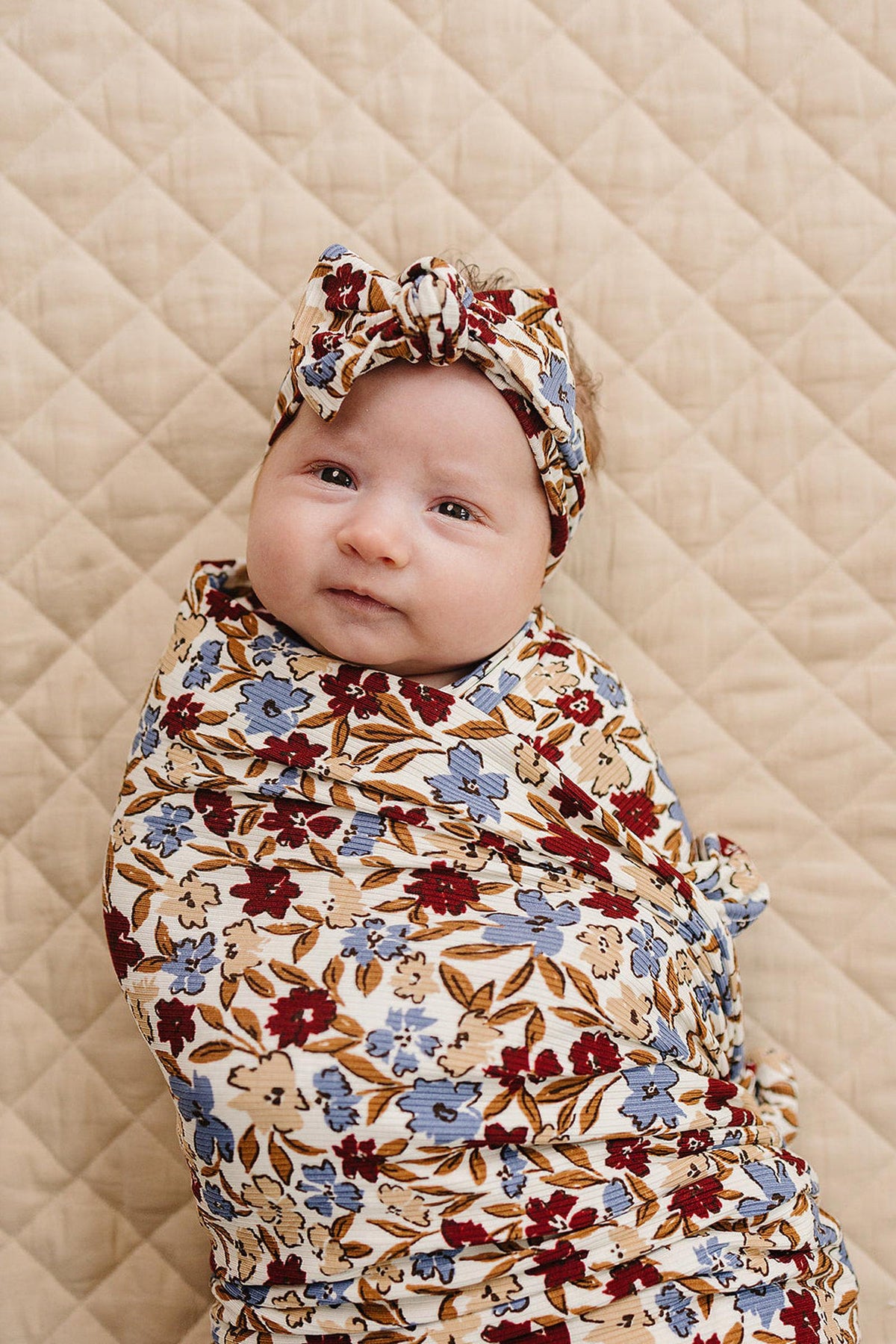 A baby wrapped in a floral-patterned head wrap and swaddle, lying on a quilted beige background.