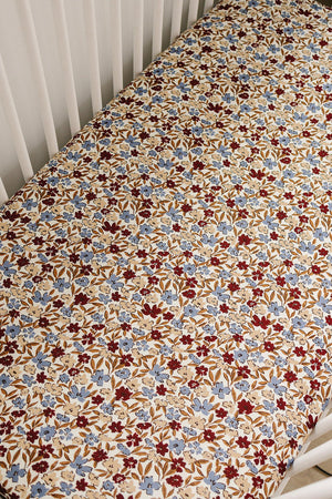 Colorful floral-patterned crib sheet with blue and burgundy flowers on a beige background, fitted in a white crib.