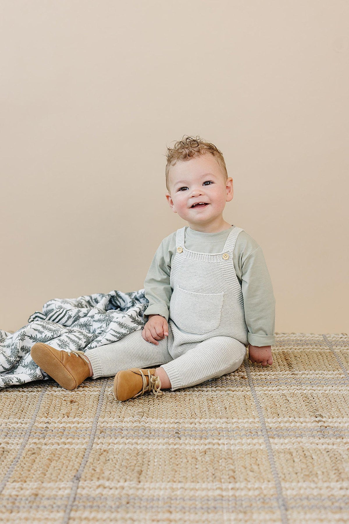 A smiling baby wearing grey knit overalls and brown shoes sits on a woven mat with a patterned blanket.
