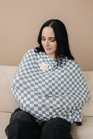 A woman sits on a beige couch, using a blue checkered bamboo cover with a Mebie Baby label.