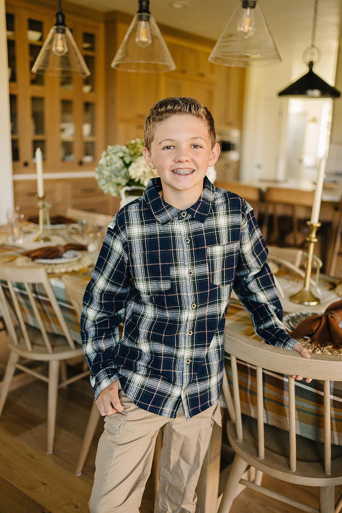 A young boy stands smiling in a cozy kitchen, wearing a navy plaid button-up shirt made from soft cotton. The shirt features a chest pocket and a classic collar.