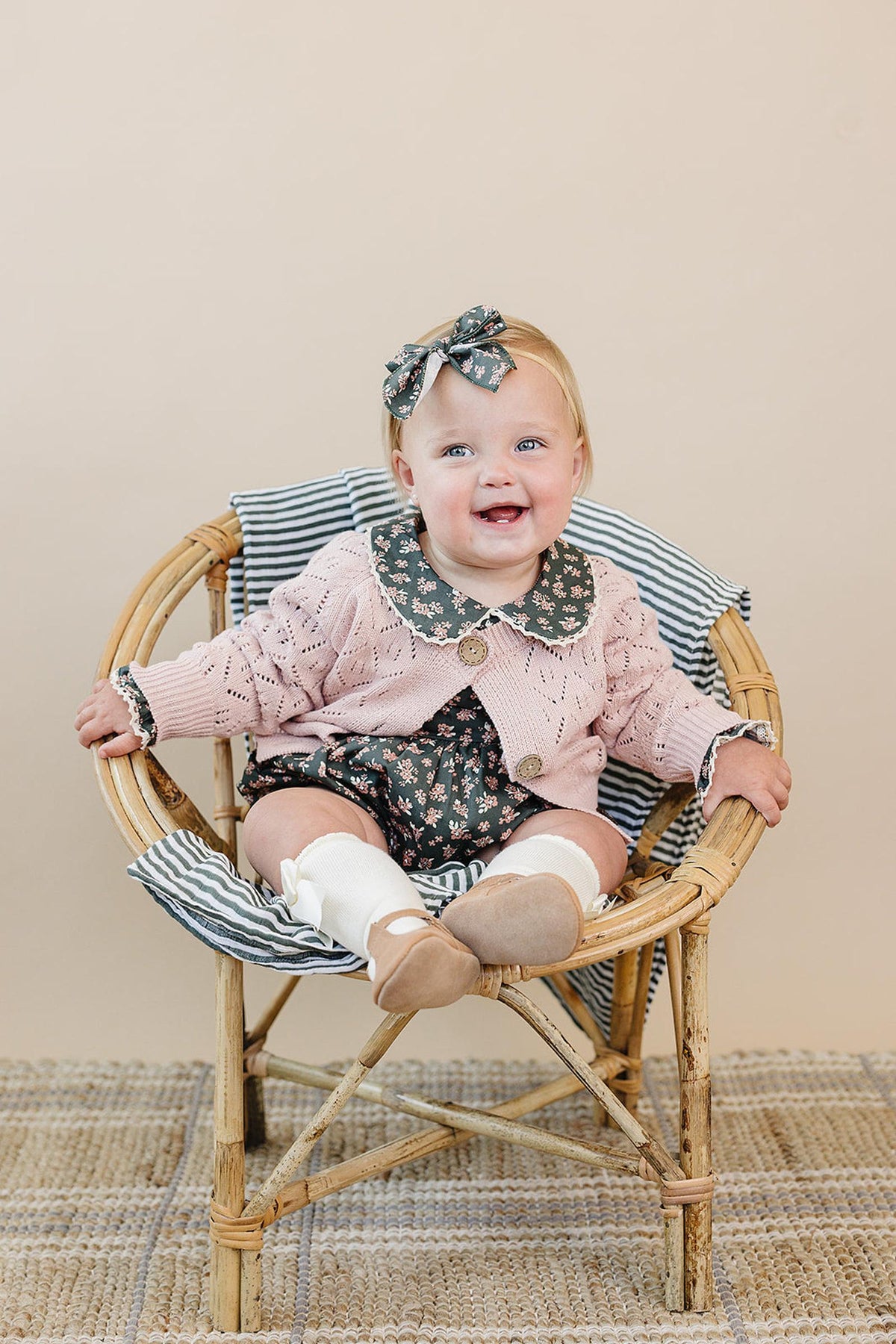 A baby wearing a pink pointelle cardigan and floral outfit sits in a wooden chair with striped cushion.