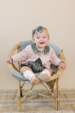 A baby wearing a pink pointelle cardigan and floral outfit sits in a wooden chair with striped cushion.