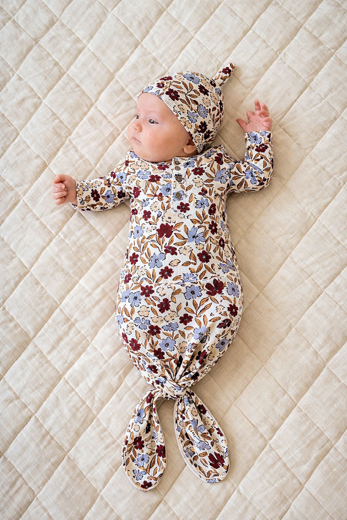A newborn lies on a quilt, wearing a floral-patterned bamboo knot gown and matching hat, showcasing soft, sustainable fabric.