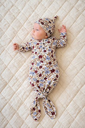 A newborn lies on a quilt, wearing a floral-patterned bamboo knot gown and matching hat, showcasing soft, sustainable fabric.