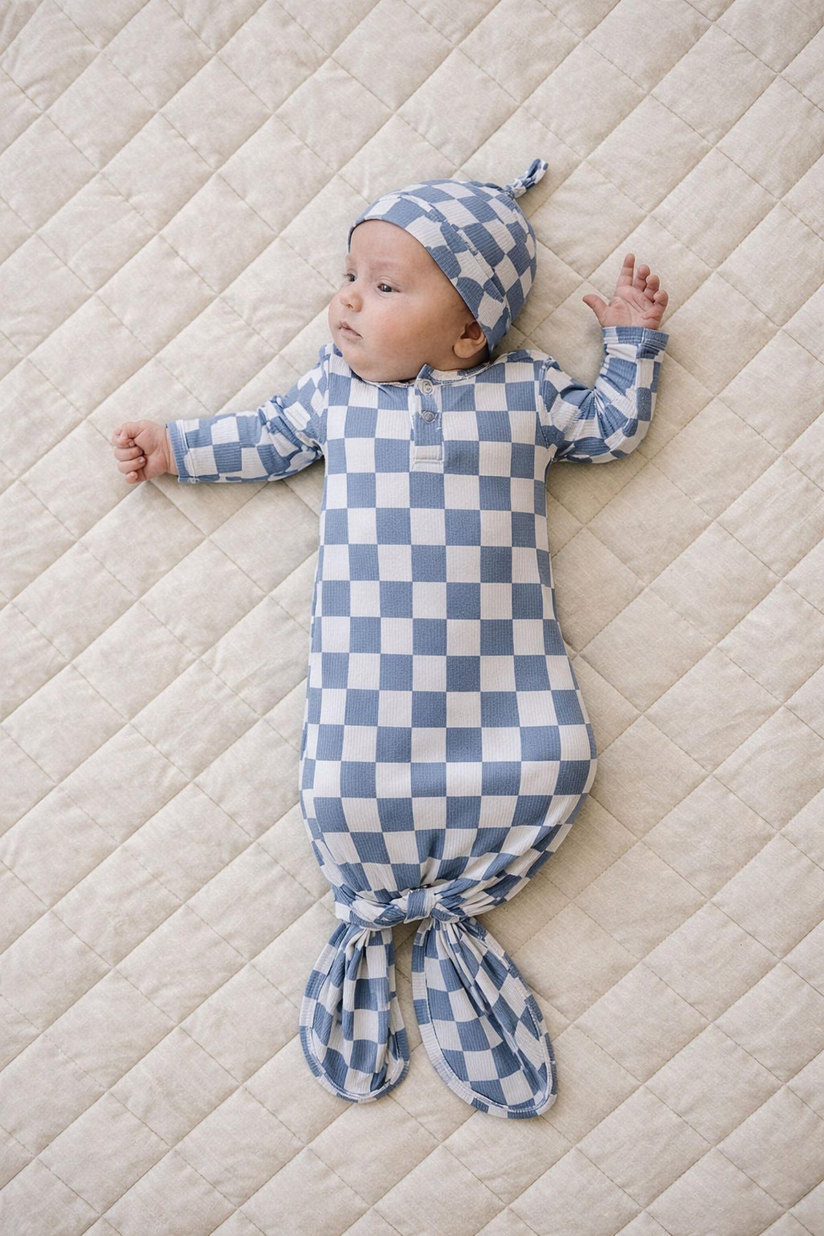 A baby in a blue checkered ribbed bamboo knot gown and matching hat lies on a cream quilted surface.