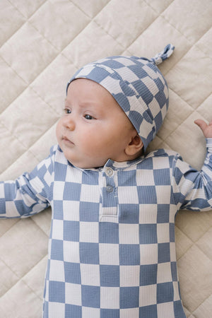 A baby lies on a quilted surface, wearing a blue and white checkered outfit and a matching knot hat.