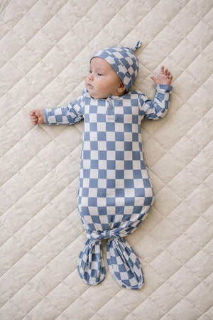 A baby in a blue checkered ribbed bamboo knot gown and matching hat lies on a cream quilted surface.