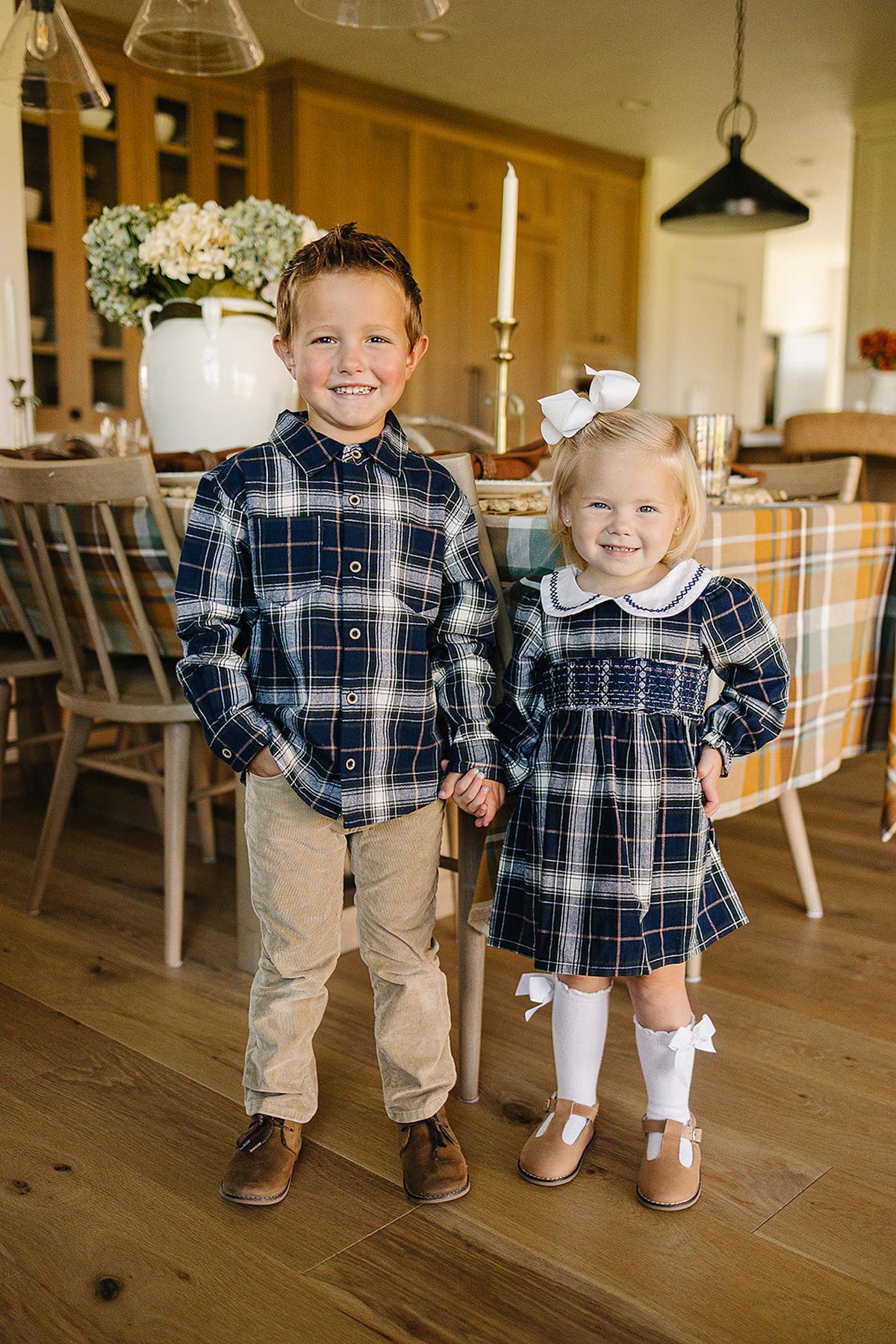 Two young children stand together in a cozy dining area, wearing a navy plaid button-up shirt and a matching dress.