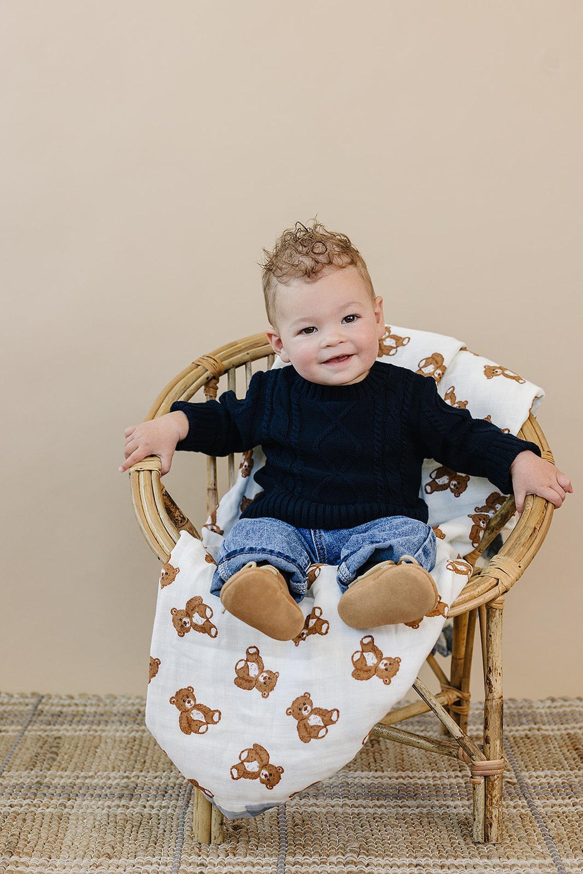 A baby in a navy cable knit sweater sits on a wicker chair with a teddy bear blanket.