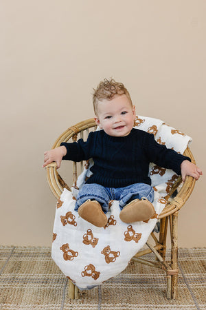 A baby in a navy cable knit sweater sits on a wicker chair with a teddy bear blanket.
