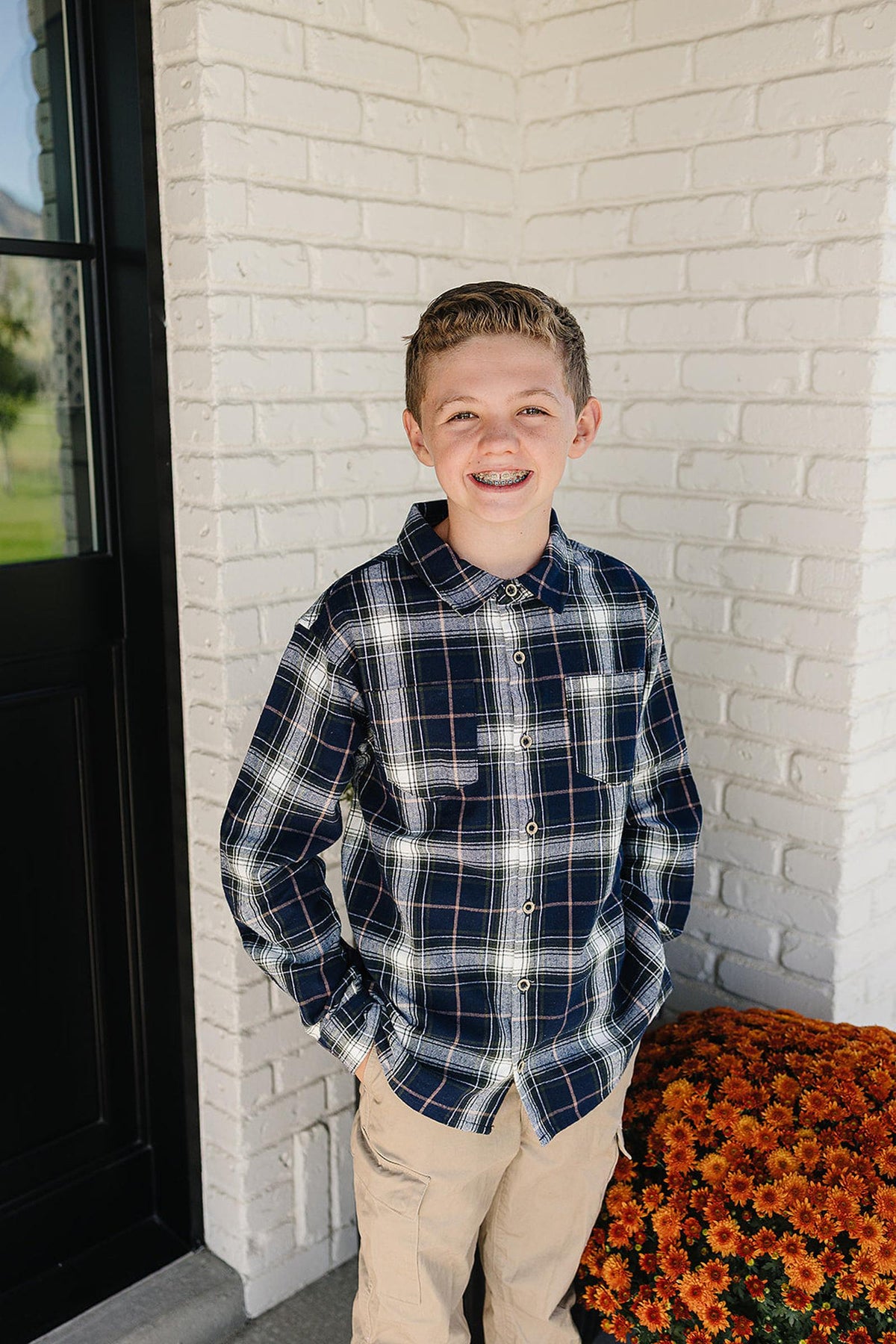 A smiling boy stands outside a white brick house, wearing a navy plaid button-up shirt made of soft cotton.