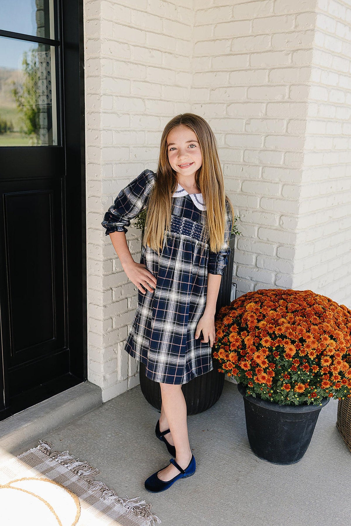A girl poses in a navy plaid smocked dress with a Peter Pan collar, standing beside a large orange flower.