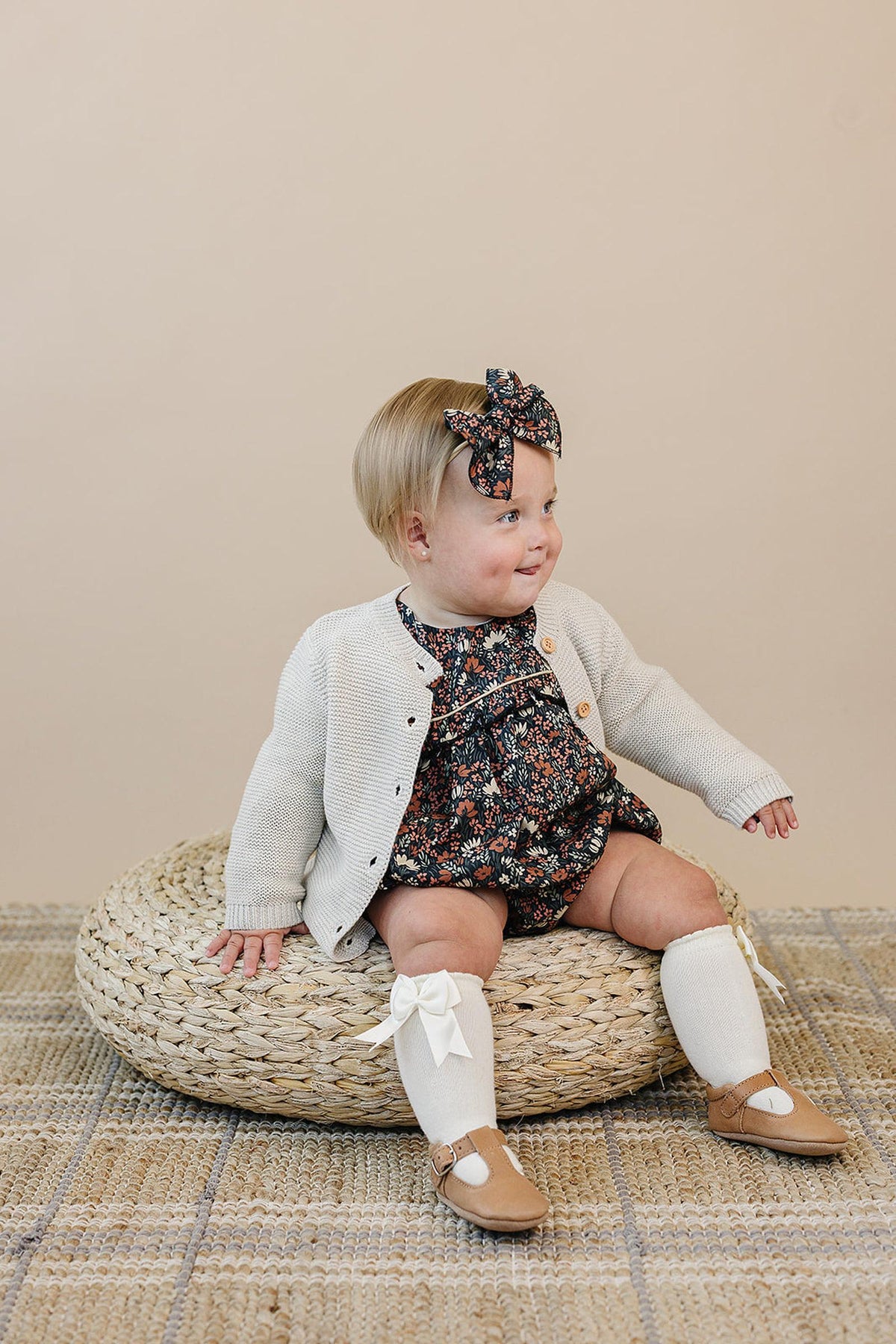 A toddler in a floral outfit and bow sits on a woven cushion, wearing a white cardigan and knee socks.