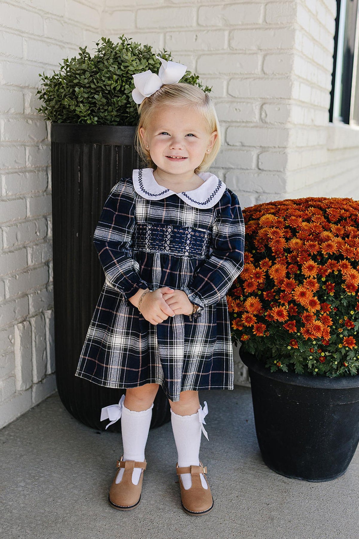 A smiling young girl wears a navy plaid smocked dress with a white Peter Pan collar, standing beside vibrant orange.
