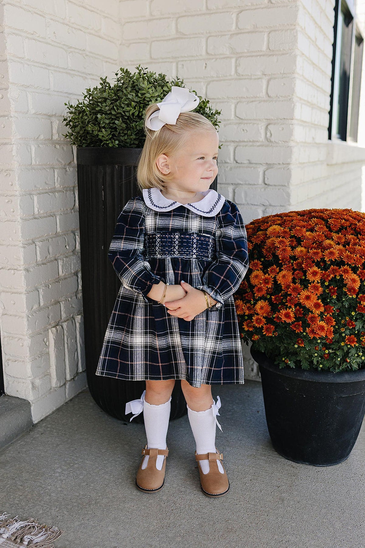 A young child stands outdoors wearing a navy plaid smocked dress with a white Peter Pan collar and brown shoes.