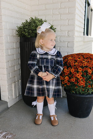 A young child stands outdoors wearing a navy plaid smocked dress with a white Peter Pan collar and brown shoes.