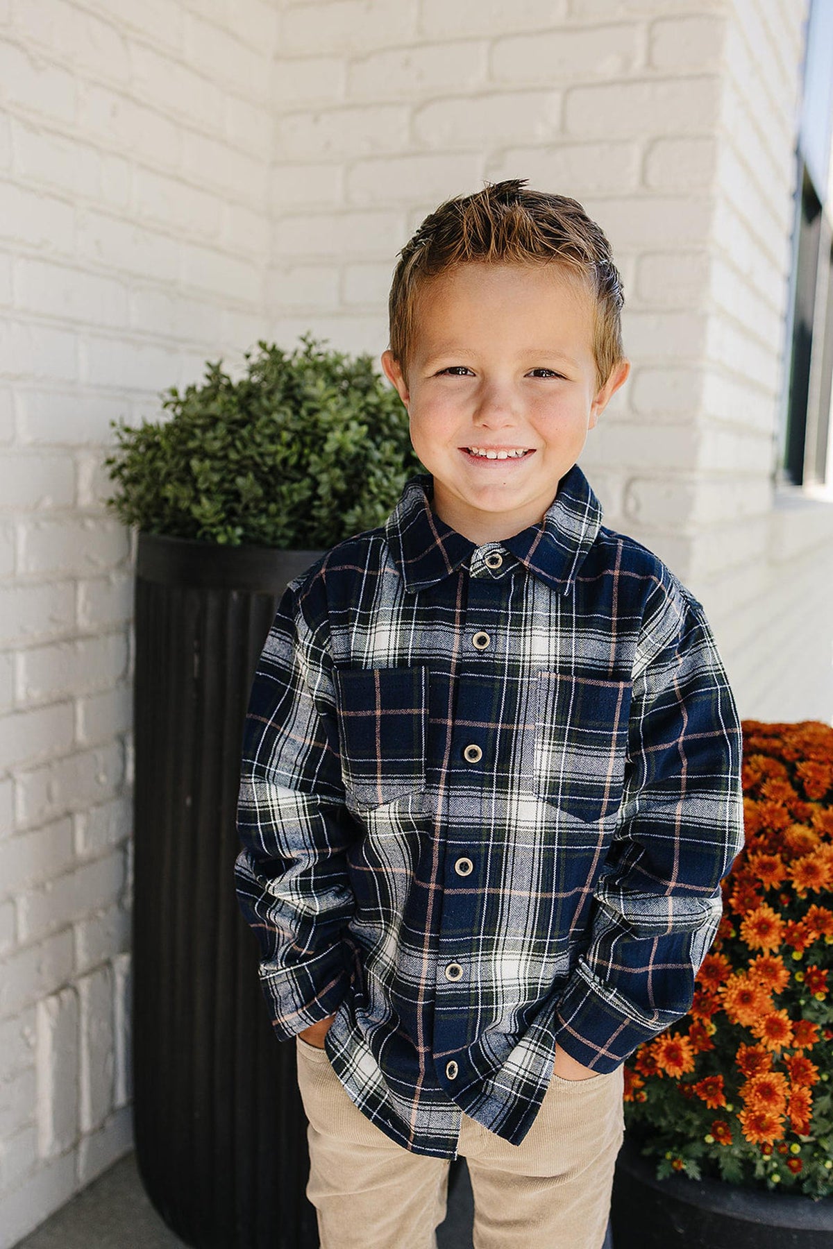 A young boy smiles while wearing a navy plaid button-up shirt made of soft cotton, standing in front of a brick wall and vibrant orange flowers.